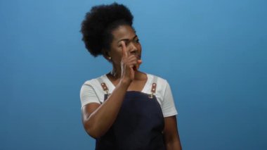 Önlük takan kadın mavi arka planda özgüvenli bir şekilde odak ve yönünü izole bir ortamda somutlaştırıyor, iddialı hareketlerle fikirleri işaret ediyor..