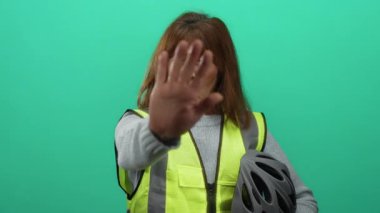 Güvenlik yeleği giyen bir kadın miğfer tutuyor ve hareketler canlı yeşil bir arkaplan karşısında duruyor, dikkati ve güvenlik bilincini odaklanmış bir ifadeyle vurguluyor..