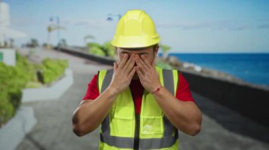 Deniz kenarında gezinen bir adam inşaat şapkası ve güvenlik yeleği giyiyor. Deniz kenarında güneşli bir gökyüzü altında yorgun görünüyor. Muhtemelen işten ya da manzaranın keyfini çıkarıyor.