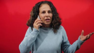Kıvırcık saçlı olgun bir İspanyol kadın telefonda canlı kırmızı bir arkaplanda konuşuyor, samimi mavi bir kazakla duygularını ifade ediyor..