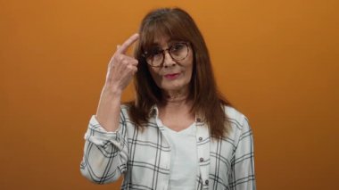 Gözlüklü kıdemli İspanyol kadın turuncu duvara başını gösteriyor, düşünceli bir ifade ve içgörü sergiliyor..