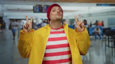 Sarı ceketli ve kırmızı bereli genç adam yoğun bir havaalanı terminalinde barış işareti yapıyor..