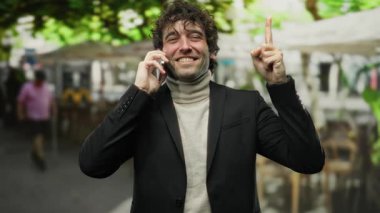 Takım elbiseli genç adam telefonda konuşurken gülümsüyor, canlı bir şehir terasında, kendine güveni ve iyimserliği somutlaştırıyor..