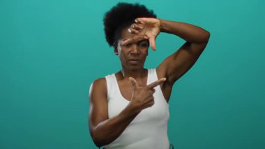 İzole edilmiş yeşil arka plana karşı elleri çerçeveli gülümseyen beyaz kolsuz bluz giyen, canlı stüdyo ortamında yakalanan neşeli hareketler yapan bir kadın..