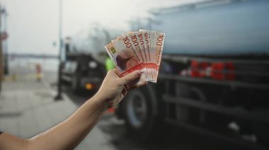Arka planda kamyonlar olan bir şehir caddesinde elinde Norveç kronu banknotları olan bir adam para ve günlük hayatı gözler önüne seriyor..