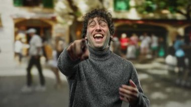 Meşgul bir şehir caddesinde gülen ve parmakla işaret eden genç adam, arka planda çeşitli kalabalıklarla canlı bir açık hava anı yakalıyor, canlı şehir hayatı sergiliyor..