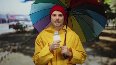 Açık havada, renkli şemsiyeli, sarı yağmurluklu ve gündüz vakti mikrofon tutan bir adam..