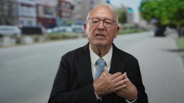 Bir sokakta kıdemli bir adam sevinçle el kol hareketleri ile açık şehir ortamında heyecan ifade ediyor, arka planda bulanık şehir manzarası ile.