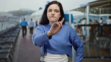 Esmer bir kadın yolcu gemisinin güvertesinde, arka planda bir sürü insan ve şezlongla el kol hareketi yapıyor.