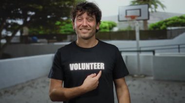 İspanyol adam gülümsüyor ve açık hava basketbol sahasında gönüllü gömleğe işaret ediyor, açık hava ortamının kurulmasında toplumsal rol oynuyor..