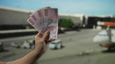 Elinde Türk lira banknotları tutan adam havaalanı terminalinde arka planda uçakla finans ve seyahat kavramını resmediyor..