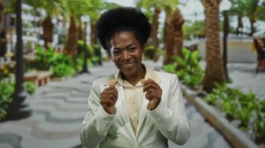 Güneşli bir açık hava parkı ortamında neşeyle dans eden bir kadın, şık beyaz bir takım elbise giyiyor, yemyeşil ve palmiye ağaçları ile çevrili, mutluluk ve enerjiyi somutlaştırıyor..