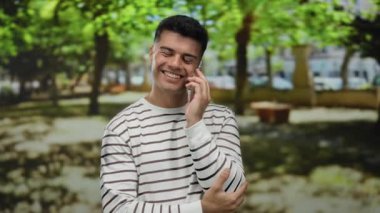 Güneşli bir park ortamında telefonla konuşan genç bir adam, çizgili bir gömlek giyiyor, doğa ile çevrili, mutlu bir ifadeyle..