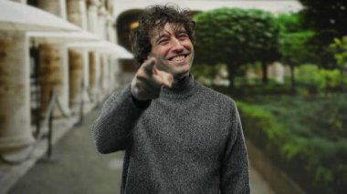 Genç adam gülümsüyor ve arka planda yeşillik ile bir şehir caddesinde işaret ediyor, rahat bir kazak giyiyor ve büyüleyici bir gülümseme ile cana yakın ve neşeli bir titreşim yayıyor..