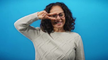 Kıvırcık saçlı, gülümseyen ve mavi arka planda poz veren, gözlük ve süveter takan, neşe ve kendine güveni ifade eden olgun bir kadın..