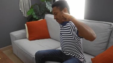 Çizgili gömlekli genç adam rahat bir oturma odasındaki koltukta rahatsiz bir sekilde geriniyor..