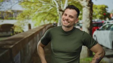 Arka planda ağaçlar ve arabalarla güvenli bir şekilde gülümseyen genç adam..