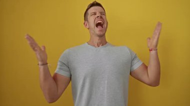 Genç adam coşkuyla sevinçle bağırıyor, parlak sarı bir arka planda, günlük gri tişört giyiyor ve kollarını açıp heyecanını dile getiriyor..