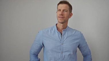 Mavi gömlekli, beyaz duvara karşı güvenle gülümseyen genç adam, rahat ve çekici bir görünüm sergiliyor..