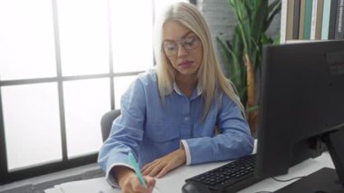 Modern bir ofiste çalışan bir kadın, bilgisayarlı bir masada otururken notlar yazmaya odaklanmış, doğal ışık ve bitkilerle çevrili, gözlük takıyor ve günlük giysiler giyiyor..