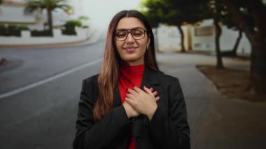 Gözlüklü bir kadın sokakta dikiliyor. Siyah bir ceket giyiyor ve kırmızı bluzlu. Gözleri kapalı ve elleri bir arada..