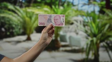 Adamın elinde Türk lirası banknotları var. Bulanık bir restoranın terasında. Arka planda yeşil bir hava var..