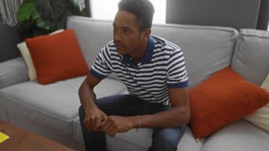 Young man celebrating in the living room with fists raised, surrounded by cozy sofa and cushions, conveying joy, indoors, casual, and relaxed ambiance.