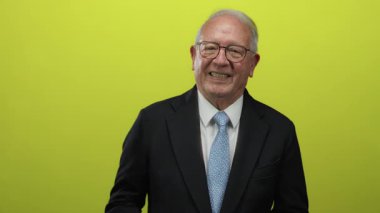 Takım elbiseli kıdemli işadamı, coşku ve pozitifliği aktaran parlak sarı arka planda el kol hareketi yapıyor..