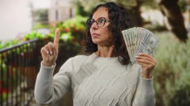 Orta yaşlı bir kadın parkta elinde Amerikan dolarlarıyla gülümseyip işaret ediyor. Bahçe geçmişiyle mali başarıyı resmediyor..