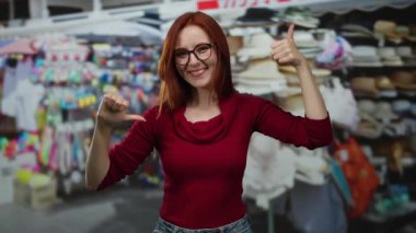 Kırmızı saçlı ve gözlüklü bir kadın, açık hava pazarında gülümsüyor, kırmızı bir kazak giyiyor, başparmaklarıyla jestler yapıyor, renkli dükkanlar ve şapkalarla çevrili..