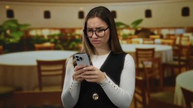 Gözlüklü genç bir kadın telefonuna şaşkın bir şekilde bakar. Kapalı bir restoranda, ahşap sandalyeler ve yuvarlak masalarla durur. Kafa karışıklığı ve merakı iletir..