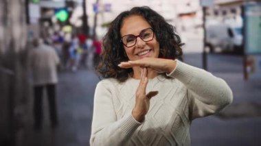 Kıvırcık saçlı, olgun bir İspanyol kadın şehir caddesinde gözlüklü ve bej kazaklı, bulanık şehir hayatıyla çevrili bir yerde mola veriyor..