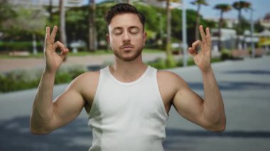 Şehir sokaklarında meditasyon yapan genç bir adam, dua eden palmiyeler, beyaz atlet giyen, bulanık yeşil ağaçlarla çevrili, sükuneti ve dışarıdaki farkındalığı ifade eden.