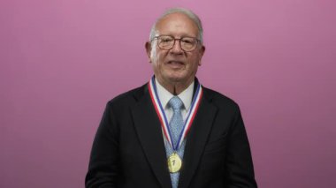 Takım elbise ve madalya giyen kıdemli bir adam pembe bir arka plana karşı ellerini kaldırarak gülümsüyor..