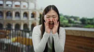 İtalya 'da Roma Kolezyumu' nun yakınında bağıran bir kadın, uzun düz saçlı ve beyaz gömlekli jestler yapıyor, açık havada canlı bir şehir ortamında..