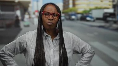 Kıvırcık saçlı bir kadın şehir caddesinde kollarını kavuşturmuş ve düşünceli bir ifadeyle etrafta binalar ve araçlarla bir şehir manzarası yakalıyor..