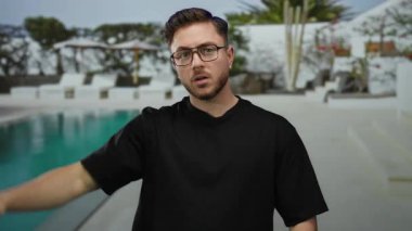 Sakallı genç bir adam otelin havuzunda, sarp bir tatil köyü bahçesiyle çevrili, siyah bir gömlek ve gözlük takıyor, saygısız bir hareket sergiliyor, rahatlık ve rahatlama vurguluyor..