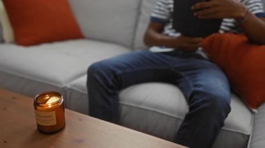 Genç adam rahat oturma odasındaki kanepede kitap okuyor turuncu yastıklar ve yanan mumlarla ılık bir atmosfer yaratıyor..