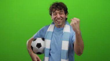 İspanyol adam mavi tişört ve eşarp giymiş futbol topuyla canlı yeşil arka plana karşı kutlama yapıyor. Heyecan ve spor coşkusu gösteriyor..