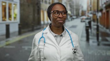 Gözlüklü ve steteskoplu kadın doktor şehir geçmişine karşı düşünceli bir ifadeyle şehir sokağında duruyor..