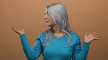 Mavi kazaklı yaşlı kadın, izole edilmiş kahverengi arka plan duvarına el hareketi yaparak rahat ve düşünceli bir davranış sergiliyor..