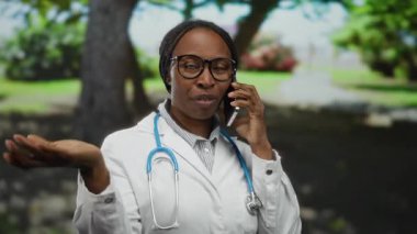 Steteskop takan ve gözlük takan kadın doktor güneşli bir park ortamında akıllı telefonla konuşuyor, dışarıda doğal ortamda profesyonel sağlık hizmetleri iletişimini özetliyor..