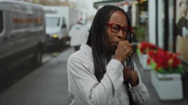 Şehir caddesinde kırmızı gözlüklü ve çizgili tişörtlü bir kadın, şehir elementleriyle çevrili, bulanık arka planlı, hareketli bir çevrede rahatsızlık anı yakalıyor..