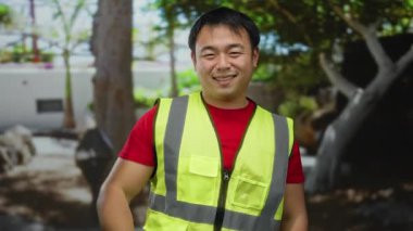 Dışarıda güneşli bir günde parkta gülümseyen güvenlik yeleği giyen genç Çinli bir adam.