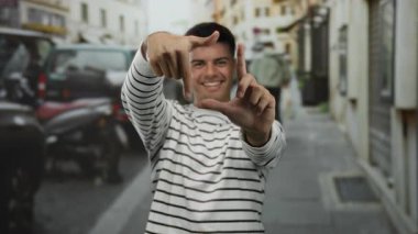 Çizgili tişörtlü genç adam gülümsüyor ve elleri ile şehir dışında canlı bir sokak arkaplanında çerçeveleniyor.