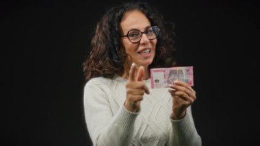 Orta yaşlı bir kadın elinde Endonezya Rupiah banknotları tutuyor ve kamerayı siyah bir duvara doğrultuyor..