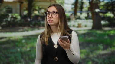 Gözlüklü genç sarışın bir kadın parkta telefona bakarken hayal kırıklığı yaşıyor. Açık hava ortamında dijital rahatsızlık tasvir ediyor..