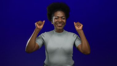 Kollarını kaldırarak kutlayan bir kadın, izole edilmiş mavi bir arka planda mutluluğu ve güveni ifade ediyor, genç bir Afro-Amerikan kadınını neşe içinde gösteriyor..