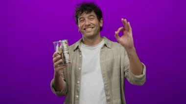 Hispanik bir adam gülümsüyor ve elinde dolarla dolu bir kumbara tutuyor canlı mor bir arka plana karşı, güven ve finansal başarıyla el kol geziyor..