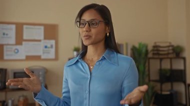 Gülümseyen bir kadın, rahat bir ofis ortamında bitkilerle ve arkada ilan panosuyla, gözlük ve mavi bir bluz takarak güven ve profesyonelliği ifade ediyor..